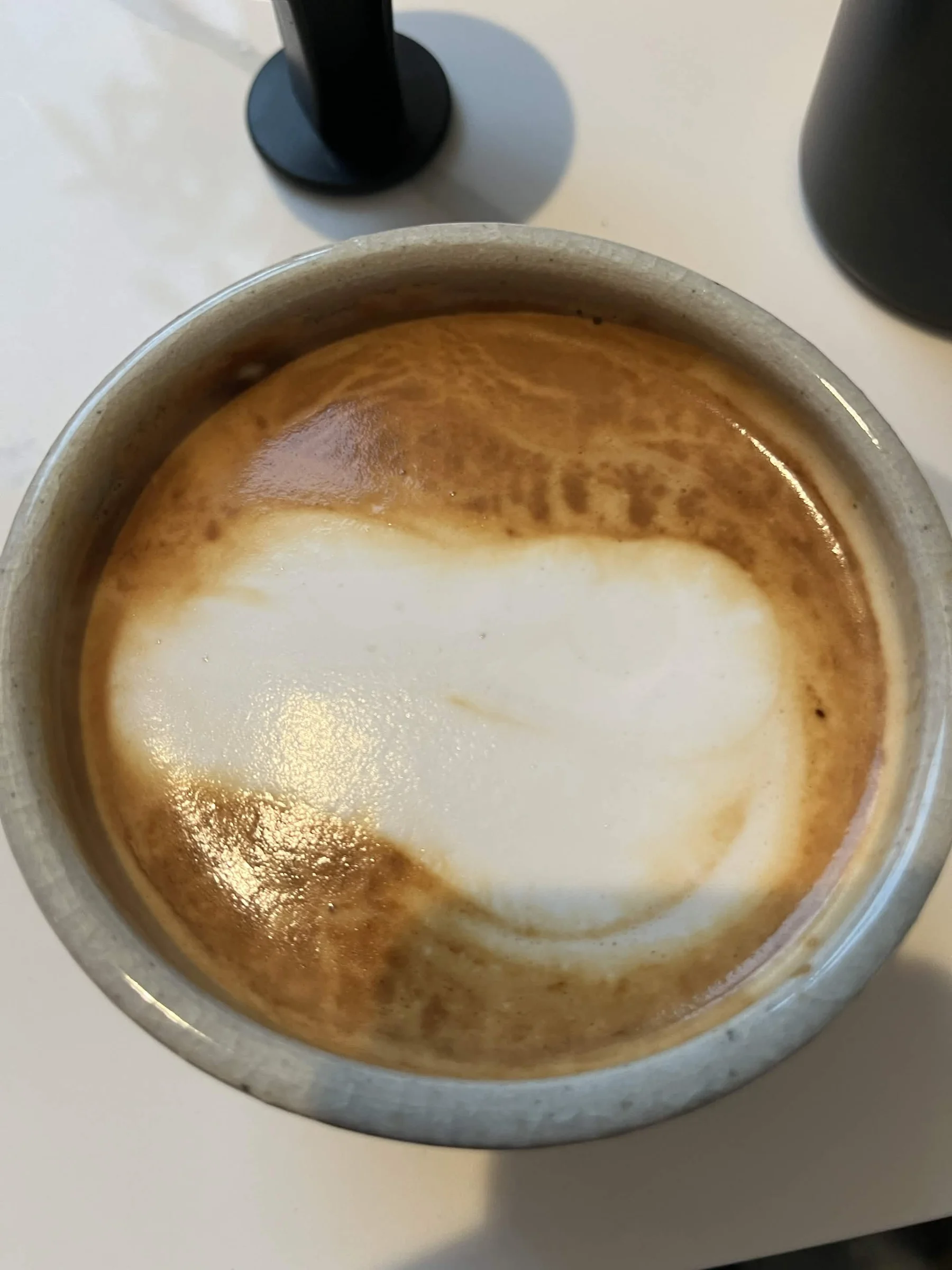 Latte in a speckled grey ceramic cup with a broad, uneven white foam shape across golden-brown crema, seen from above.
