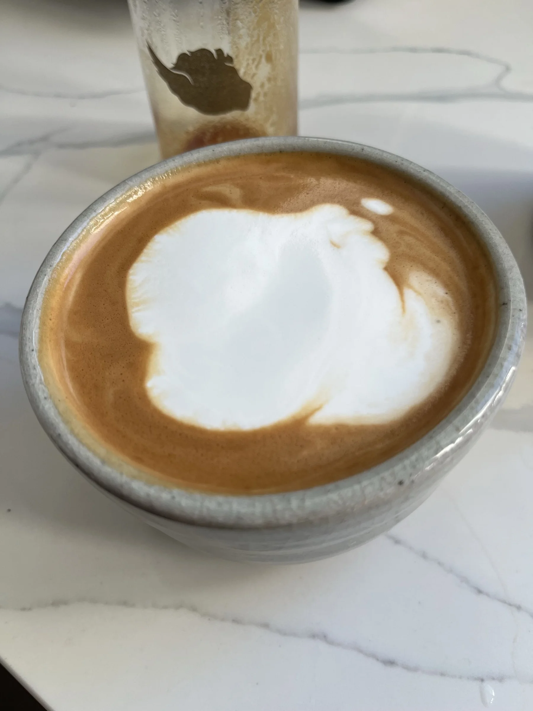 Latte in a speckled ceramic cup with a broad white foam shape and a few small foam droplets near its top edge.
